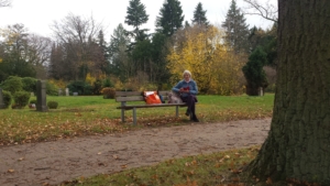 Eine Frau sitzt auf einer Bank und schaut in die Kamera. Die Bank steht in einem Park. Es ist Herbst.