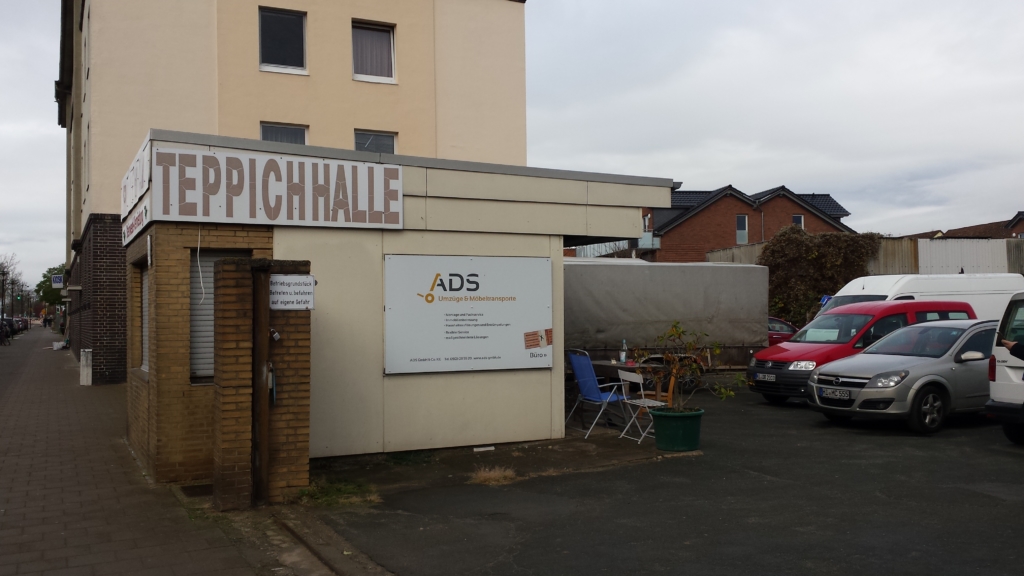 Ein Gebäude mit der Aufschrift "Teppichhalle". Rechts im Bild parken einige Autos.