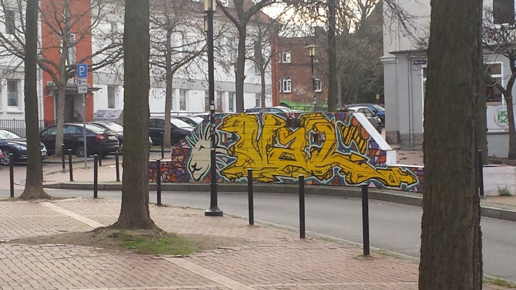 Ein Platz in Hildesheim. Im Hintergrund sind parkende Autos zu sehen. Eine Wand mit Graffities steht im Mittelpunkt.