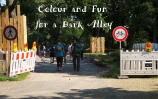 Menschen gehen einen Weg entlang. Links und rechts befindet sich eine Baustelle. Über dem Bild steht in weißer Schrift: "Colour and Fun for a Dark Alley".