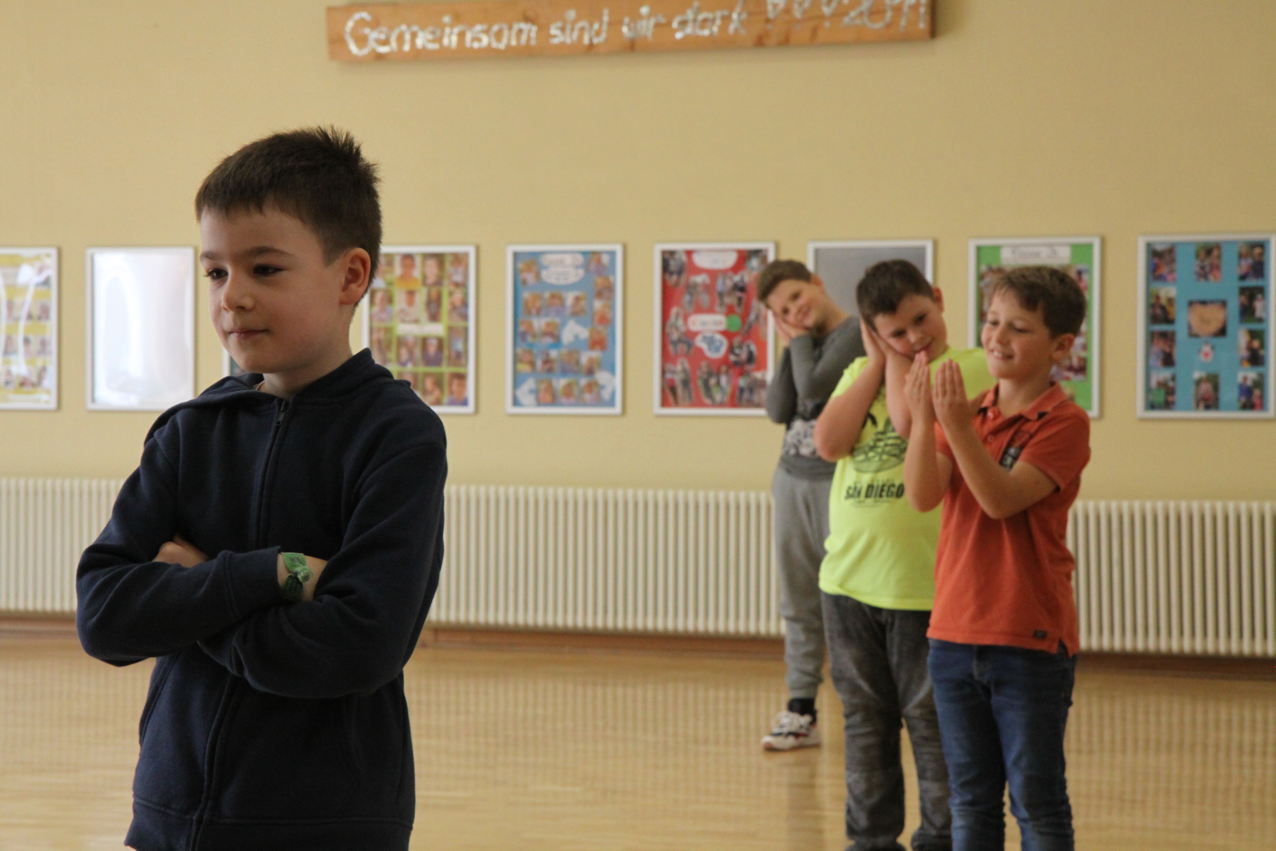 Kinder proben in der Schule ein Theaterstück. Ein Junge mit verschränkten Armen befindet sich links im Bild. Rechts stehen drei weitere Jungs und formen mit den Händen ein Kissen unter ihrem Kopf.