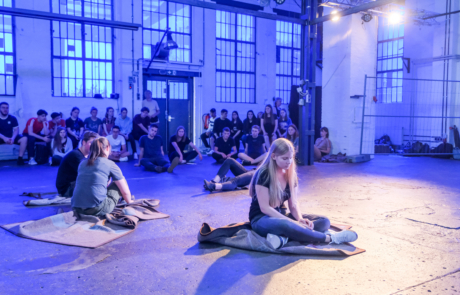 In einer blau beleuchteten Fabrikhalle sitzen Jugendliche auf Decken auf dem Boden. Im Hintergrund sitzen viele weitere Personen auf dem Boden und schauen den Jugendlichen zu.