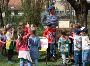 Eine Gruppe von Kindern steht auf einer Wiese. In der Mitte steht ein Mann und unterhält sich mit den Kindern.