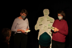 Zwei Personen stehen vor einer weißen Pappfigur. Auf der Figur kleben grüne Zettel. Die beiden Personen halten weitere grüne Zettel in den Händen.