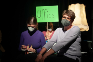 Zwei Frauen sitzen nebeneinander. Die linke Frau öffnet zwei Flaschen. Die rechte Frau lächelt. Im Hintergrund sitzt eine dritte Frau und hält ein grünes Schild hoch. Auf dem Schild steht: "Office".