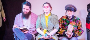 Drei Personen in Kostümen sitzen nebeneinander auf einer Bühne. Die linke Person liest ein Buch. Die rechte Person hält eine Brötchentüte in der Hand und schaut konzentriert.