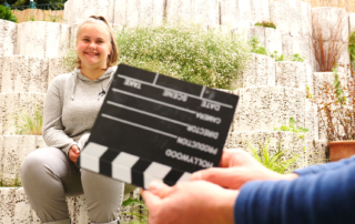 Eine junge Frau sitzt auf einem Stein und lächelt. Vor ihr hält eine Hand eine Filmklappe in die Kamera.