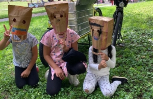 Kinder mit Papiermasken auf dem Kopf sitzen auf einer Wiese.