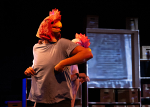 Zwei Menschen tanzen über eine Bühne. Sie tragen Hühner-Masken.