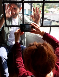 Eine Frau fotografiert einen Mann. Der Mann befindet sich hinter einem Fenster. Er drückt die Hände gegen die Fensterscheibe.