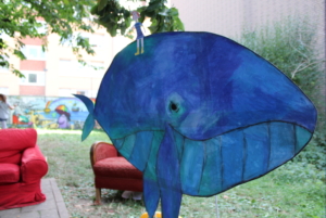 Ein großer blauer Wal aus Pappe. Auf dem Wal sitzt ein kleines Mädchen aus Papier. Im Hintergrund befinden sich zwei rote Sessel.