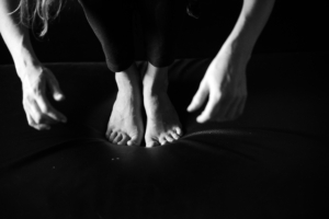 In schwarz-weiß sind die nackten Füße einer Frau zu sehen. Die Frau steht gebückt, sie lässt ihre Hände herabbaumeln.