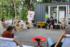 Menschen sitzen im Kreis vor einem Schiffscontainer. Eine Person liest etwas vor.