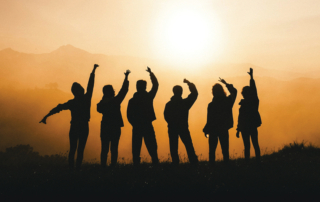 Eine Gruppe von Jugendlichen steht vor einem Sonnenaufgang. Von den Jugendlichen sind nur die Silhouetten zu sehen.