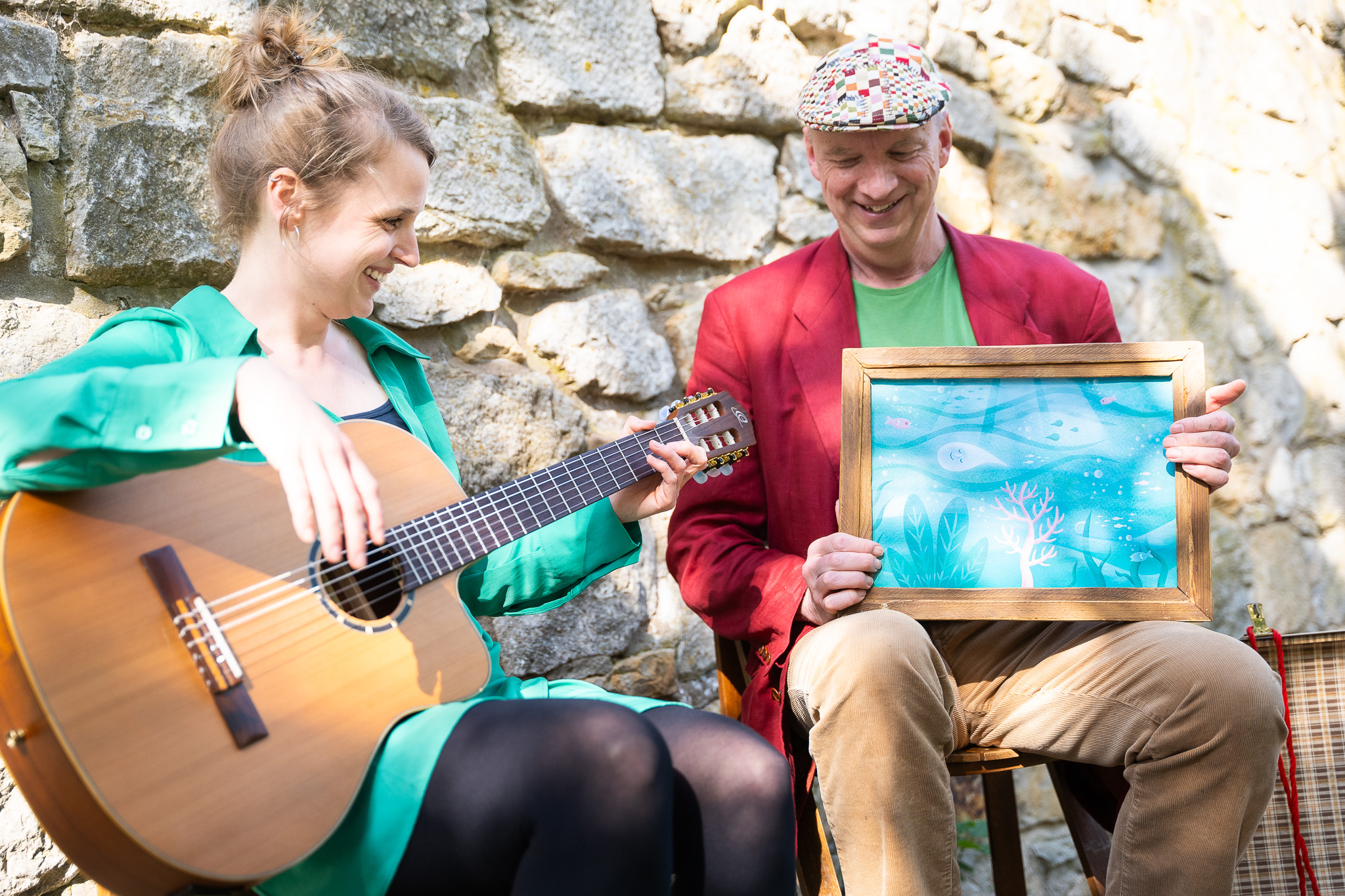 Eine Frau und ein Mann sitzen nebeneinander vor einer Mauer. Die Frau spielt Gitarre, der Mann hält ein blaues Gemälde.