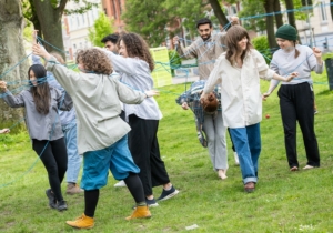 Menschen tanzen in einem Park miteinander. Durch einen blauen Faden in ihren Händen sind sie miteinander verbunden.