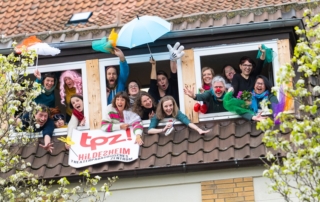 Viele verkleidete Menschen winken aus einem Fenster.