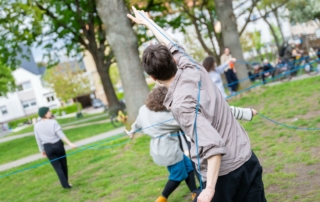 Menschen tanzen in einem Park miteinander. Sie halten einen blauen Faden in der Hand, durch den sie sich miteinander verbinden. Die Menschen sind nur von hinten zu sehen.