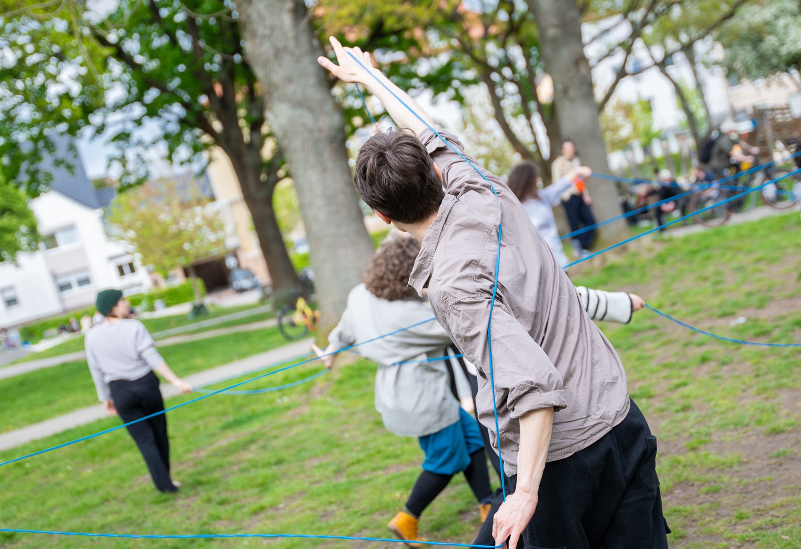 Menschen tanzen in einem Park miteinander. Sie halten einen blauen Faden in der Hand, durch den sie sich miteinander verbinden. Die Menschen sind nur von hinten zu sehen.