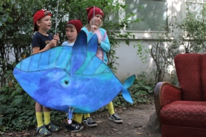 Drei Kinder stehen neben einem roten Sofa unter freiem Himmel. Sie halten einen großen, selbstgebastelten Wal in der Hand. Der Wal steht auf dem Kopf.