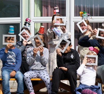 Viele Kinder sitzen gemeinsam auf einer Bank. Sie halten Bilderrahmen vor ihr Gesicht, durch die sie hindurchschauen.