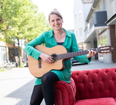 Eine Frau sitzt unter freiem Himmel auf der Ecke eines roten Sofas. Sie hält eine Gitarre in der Hand und lächelt in die Kamera.