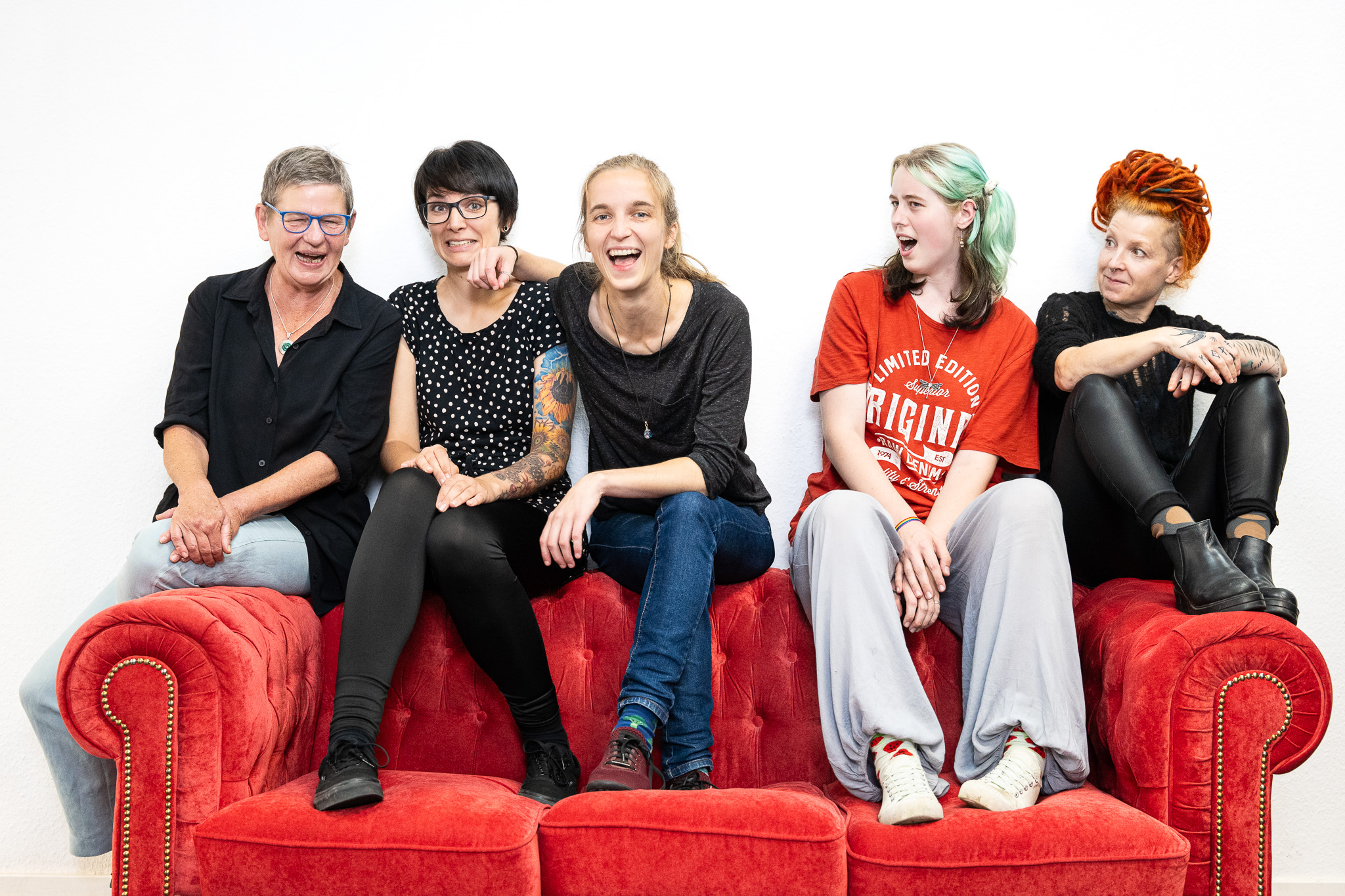 Fünf Frauen unterschiedlichen Alters sitzen auf der Lehne eines roten Sofas. Sie schneiden Grimassen.