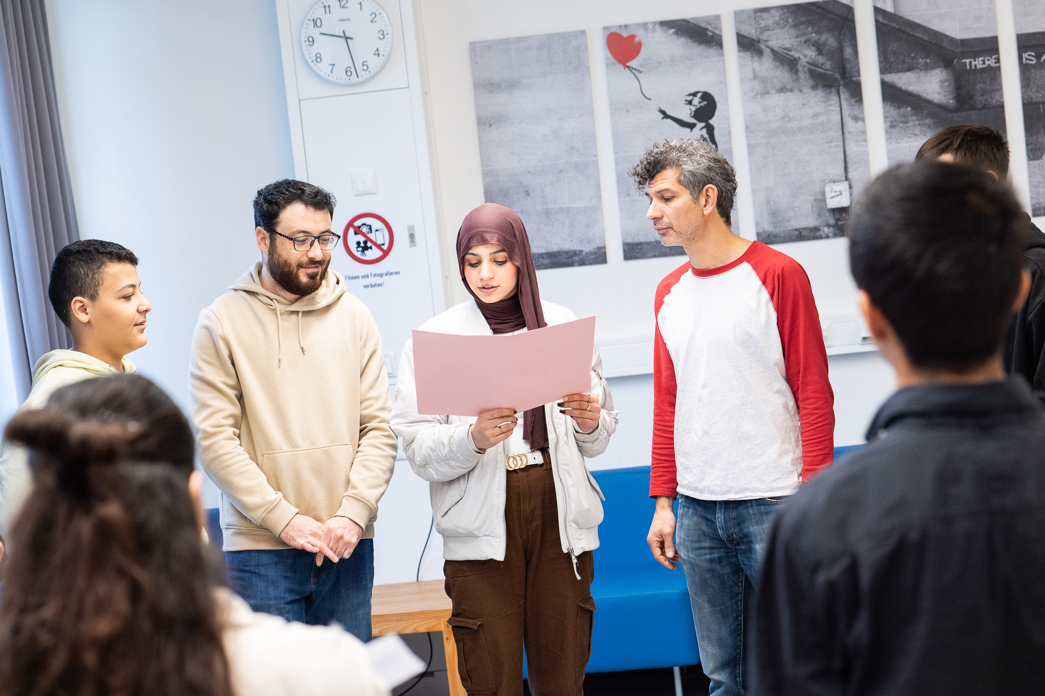 Menschen stehen zusammen in einem Klassenraum. Eine junge Frau hält einen Bogen Papier in der Hand und scheint einen Text vorzulesen.